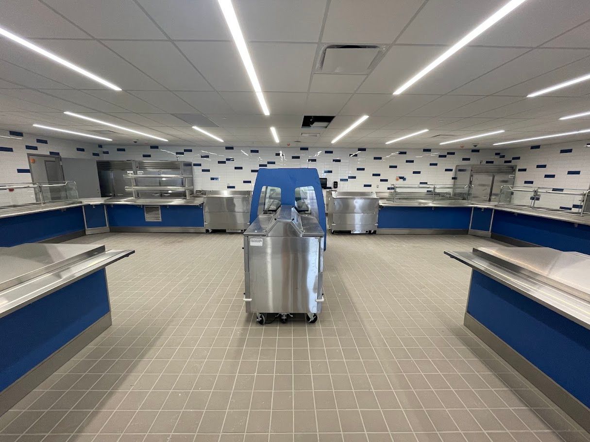 An empty cafeteria serving area featuring blue base counters, stainless steel equipment, and a patterned tiled wall.