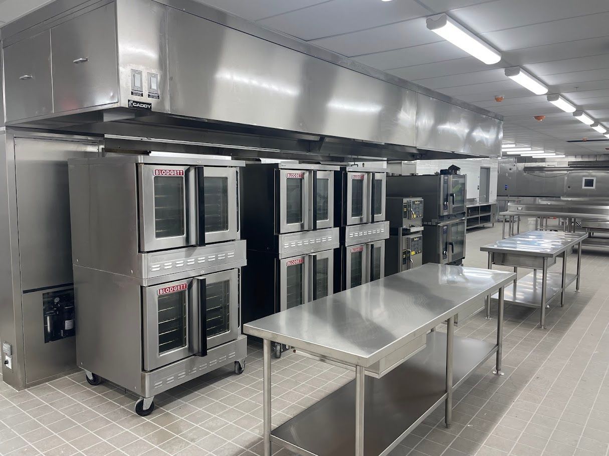 A clean, commercial kitchen with rows of stainless steel ovens under a large exhaust hood and metal prep tables.