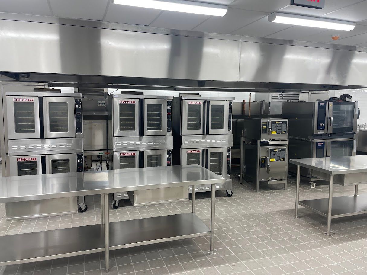 A professional kitchen with multiple stainless steel industrial ovens and workstations under a large vent hood.