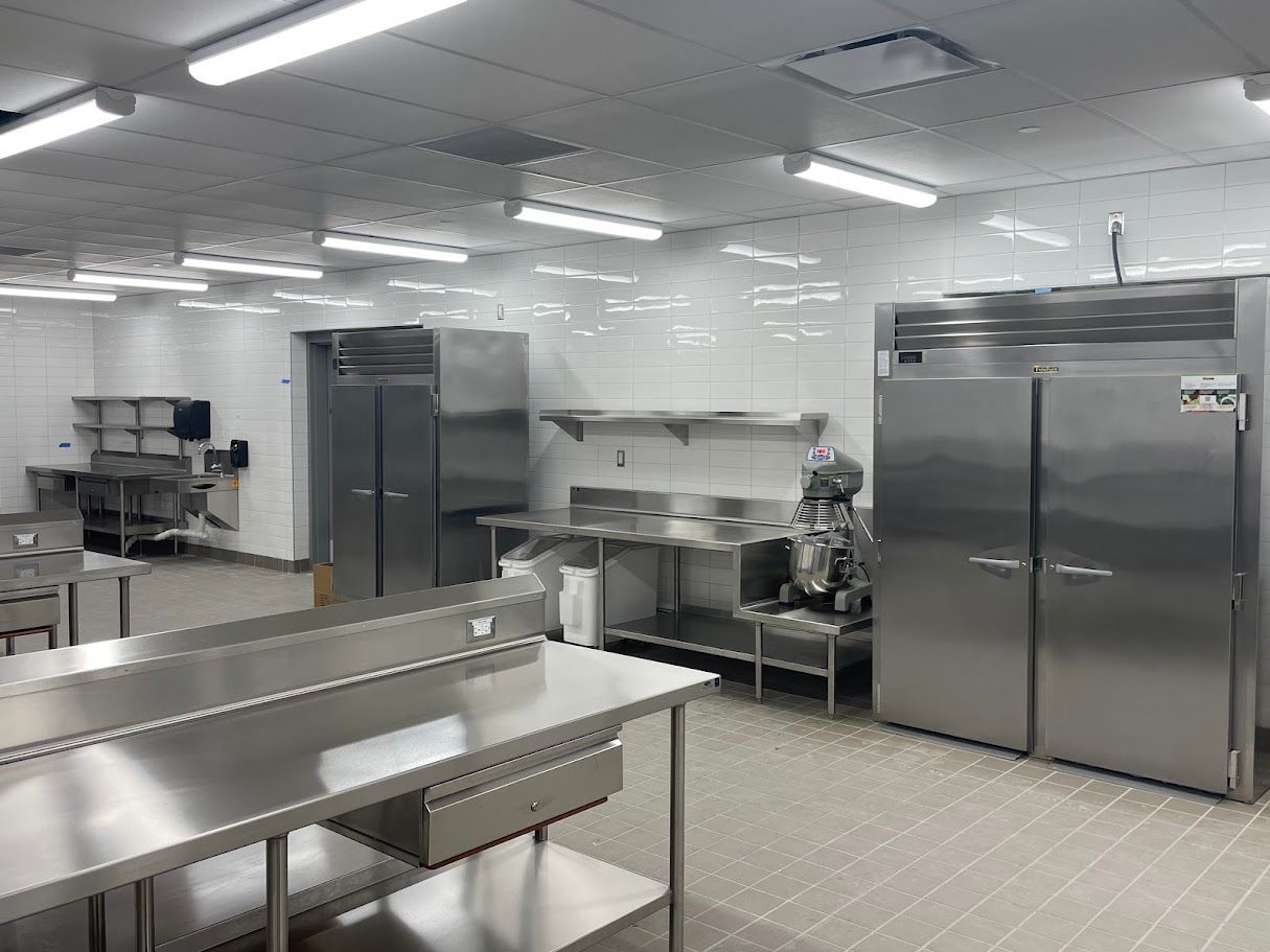 A bright, sterile commercial kitchen featuring stainless steel prep tables, large refrigerators, and white tiled walls.