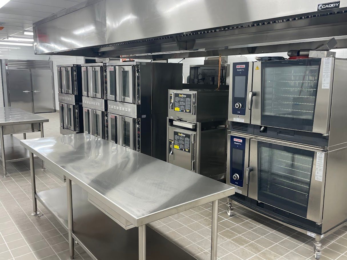 A commercial kitchen featuring stainless steel prep tables, multiple industrial ovens, and a large metal vent hood.