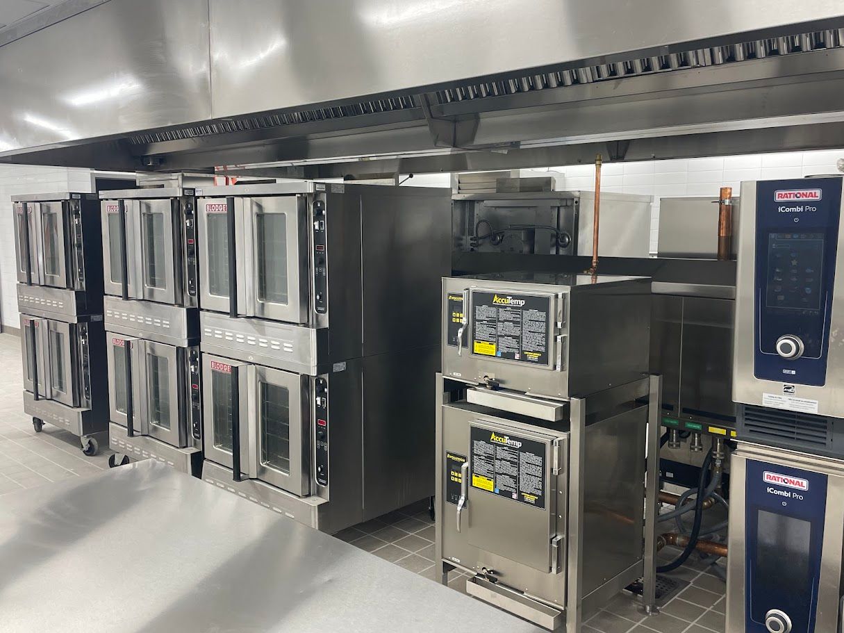 A commercial kitchen features a row of stainless steel double-stacked ovens under a large exhaust hood.