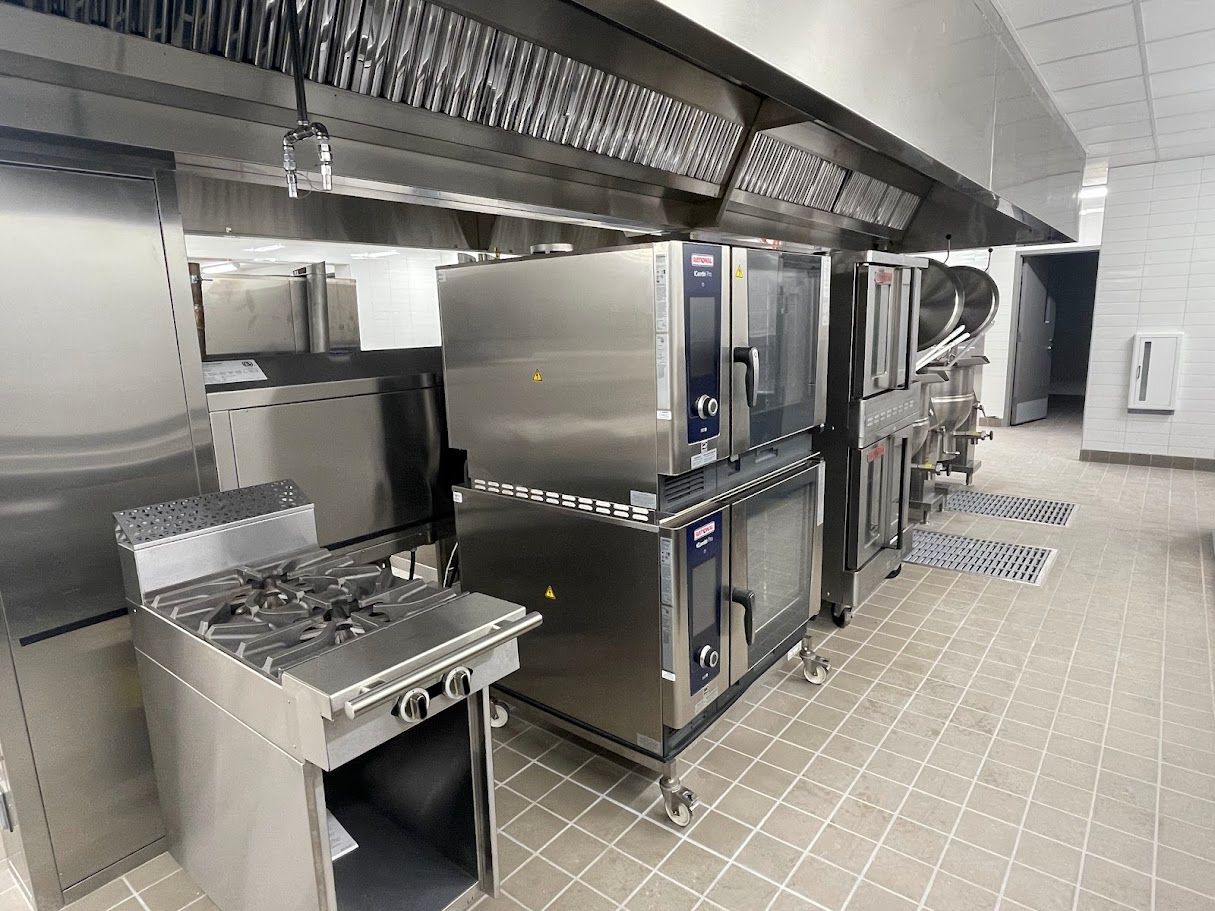 A professional kitchen with a stainless steel stove, stacked ovens, and industrial ventilation hoods on a tiled floor.