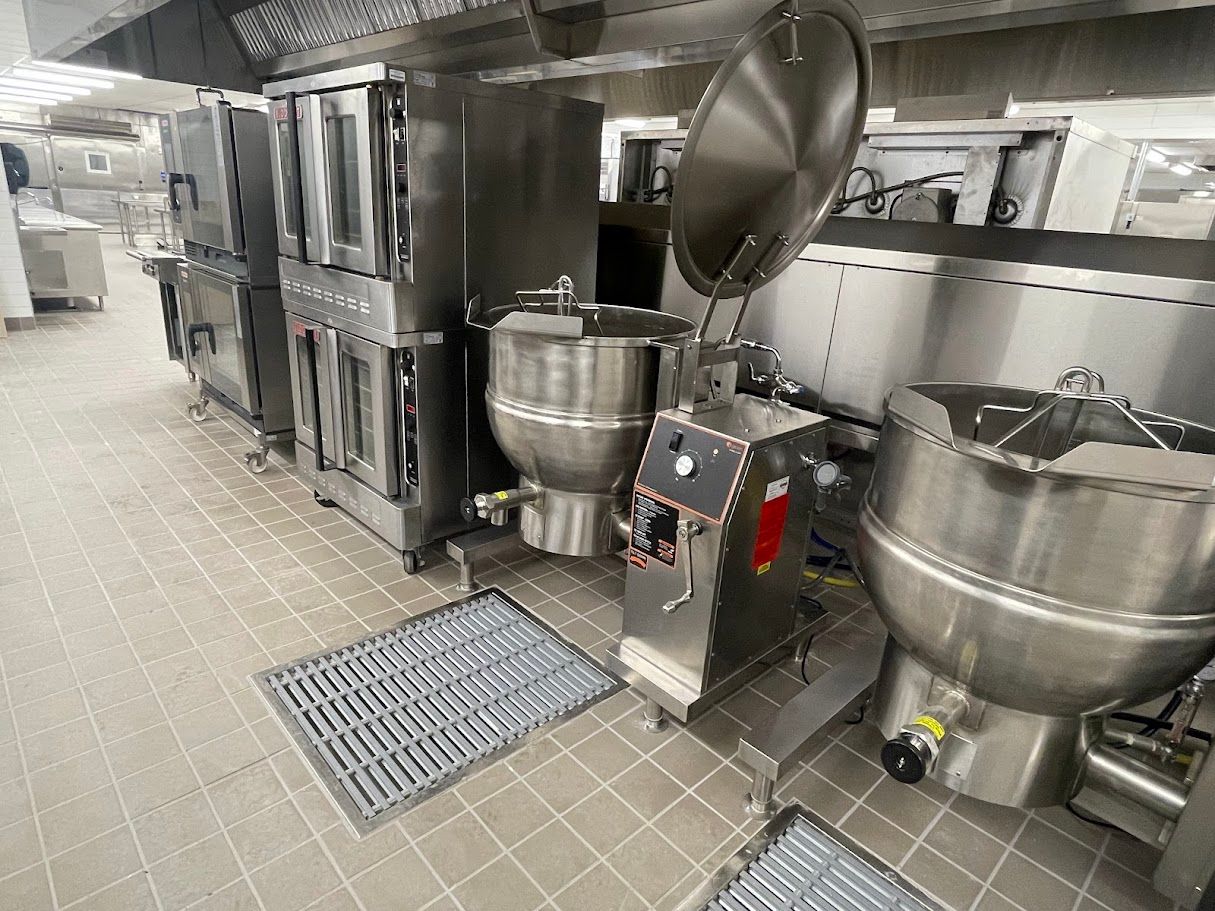 A commercial kitchen featuring stainless steel ovens and two large tilting steam kettles on a tiled floor.