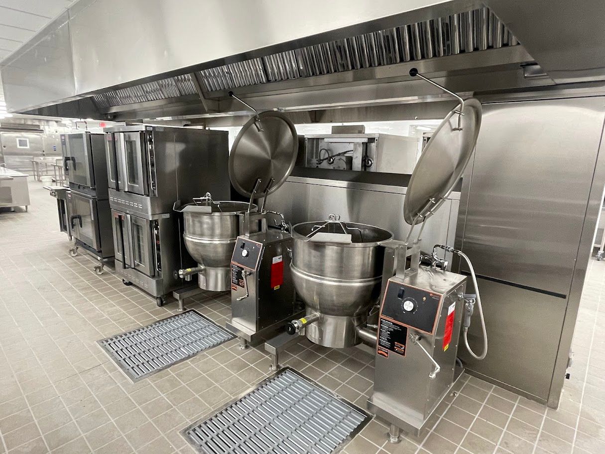 A commercial kitchen line featuring stainless steel steam kettles and stacked ovens under a large exhaust hood.