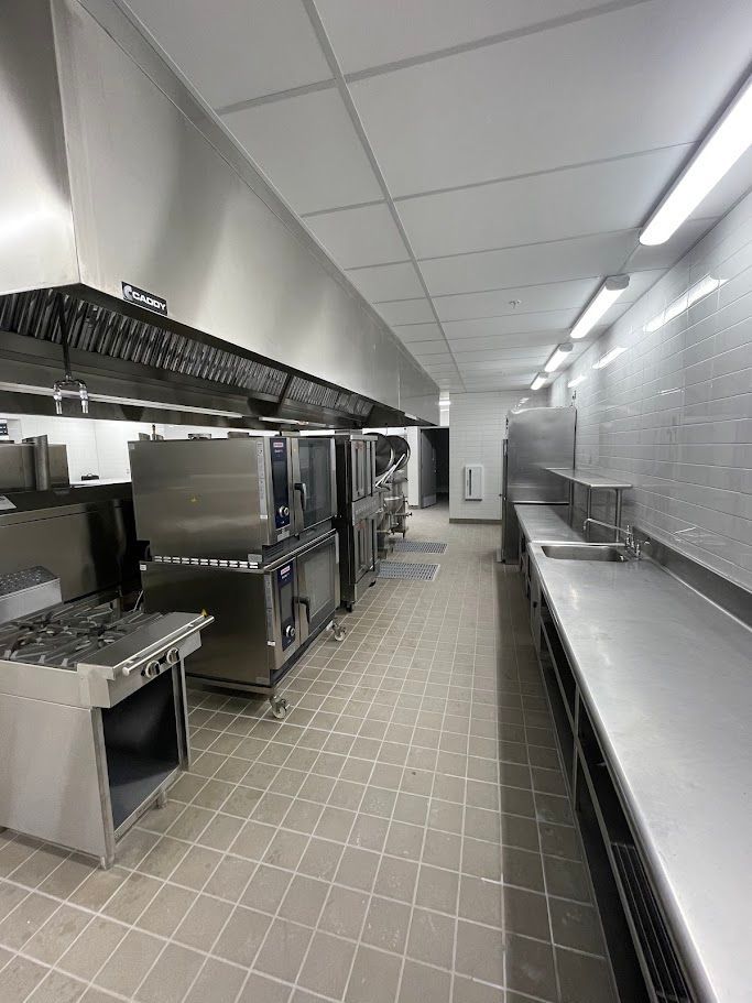 A professional kitchen featuring stainless steel stoves, ovens, a large ventilation hood, and a long preparation counter.