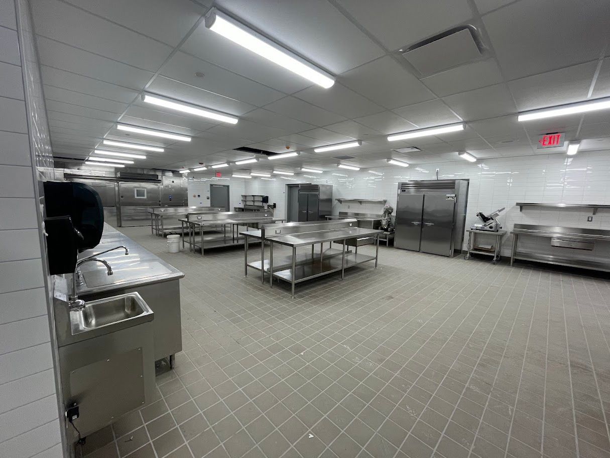 A brightly lit commercial kitchen with stainless steel prep tables, industrial appliances, and tiled walls and floors.