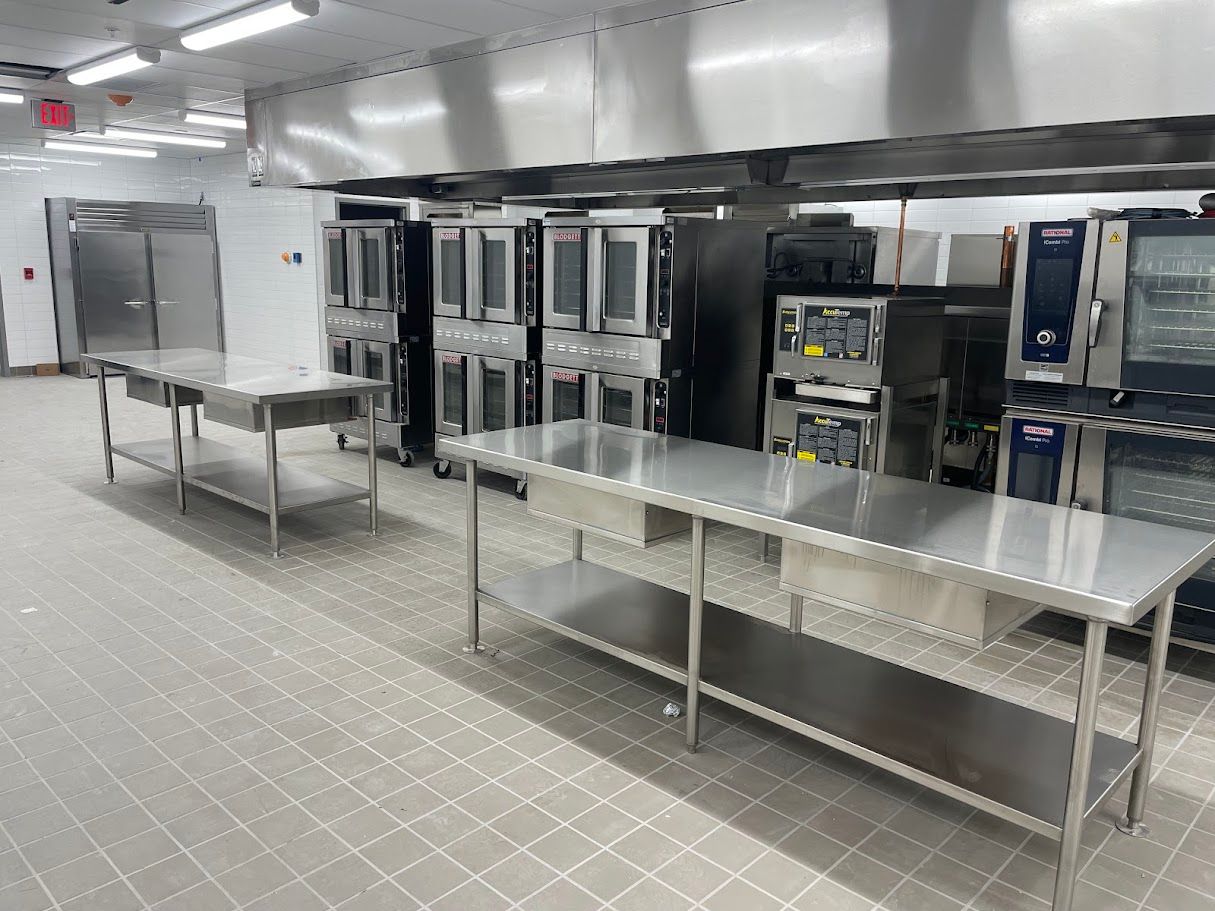Commercial kitchen with stainless steel work tables, rows of ovens, and a large ventilation hood against white walls.