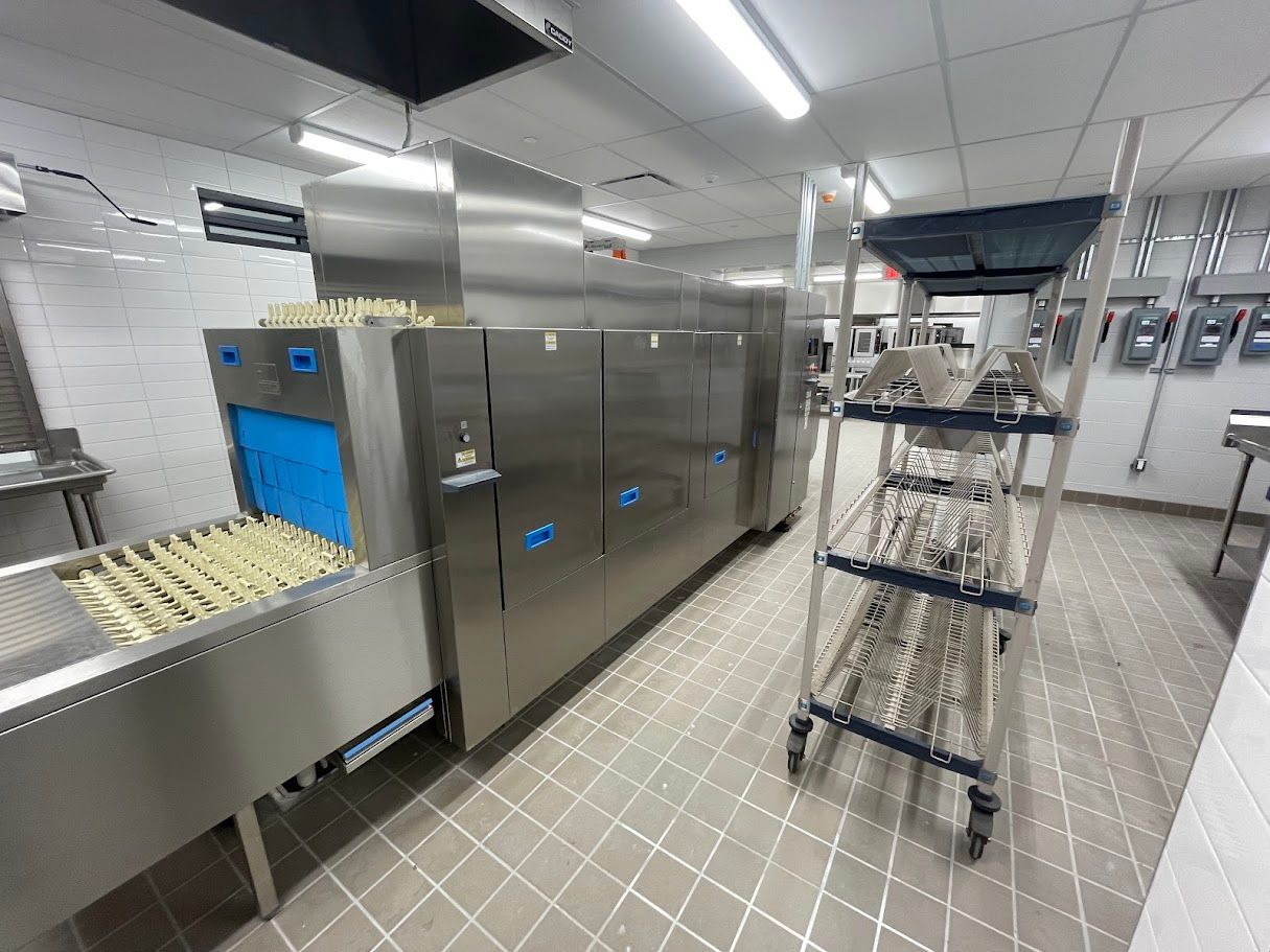 A commercial industrial dishwasher with a rack conveyor system in a tiled commercial kitchen.