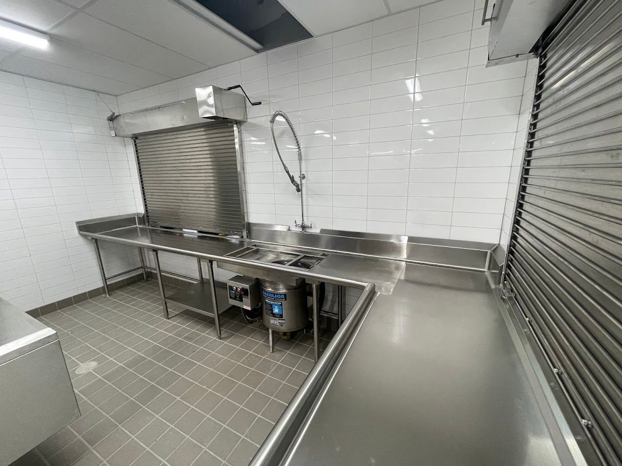 A clean, empty commercial kitchen featuring stainless steel countertops, a sink with a sprayer, and white subway-tiled walls.