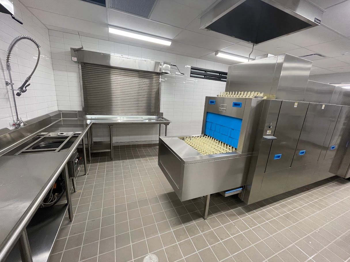 A commercial dishwashing station featuring a large, stainless steel conveyor dishwasher beside a metal prep table.