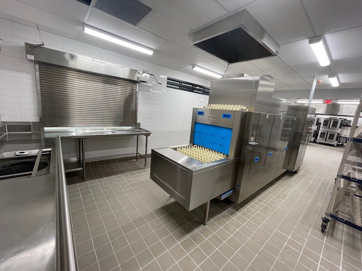 Industrial kitchen with a stainless steel commercial conveyor dishwasher cleaning trays on a tiled floor.