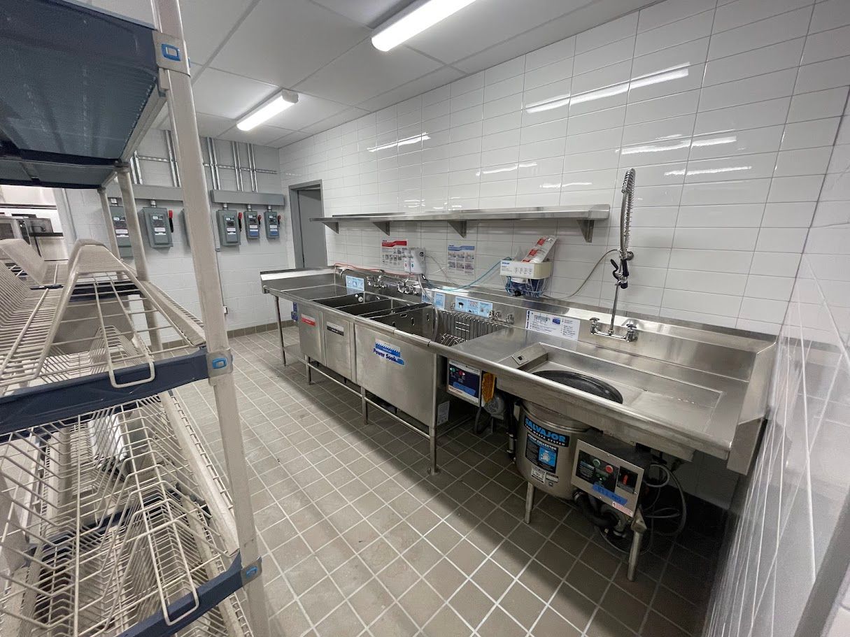 A commercial kitchen sink setup with stainless steel basins, faucets, and tiled walls, next to a wire storage rack.