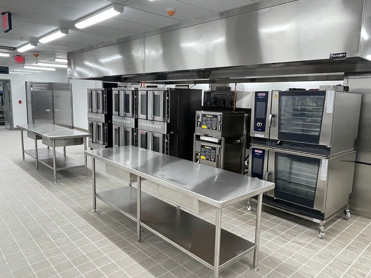 A commercial kitchen featuring stainless steel work tables, large ovens, and ventilation hoods in a tiled room.