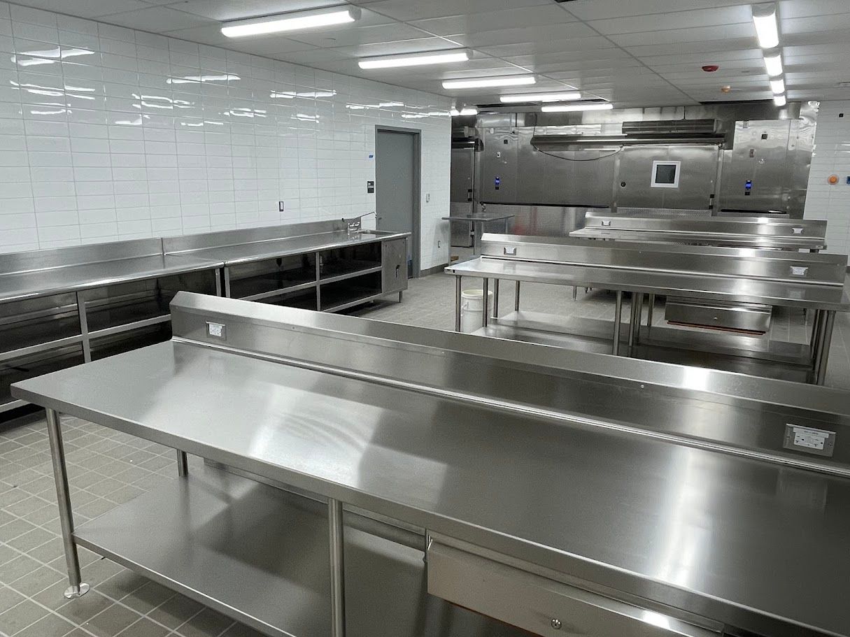 An empty commercial kitchen with stainless steel tables, white tiled walls, and bright overhead lights.