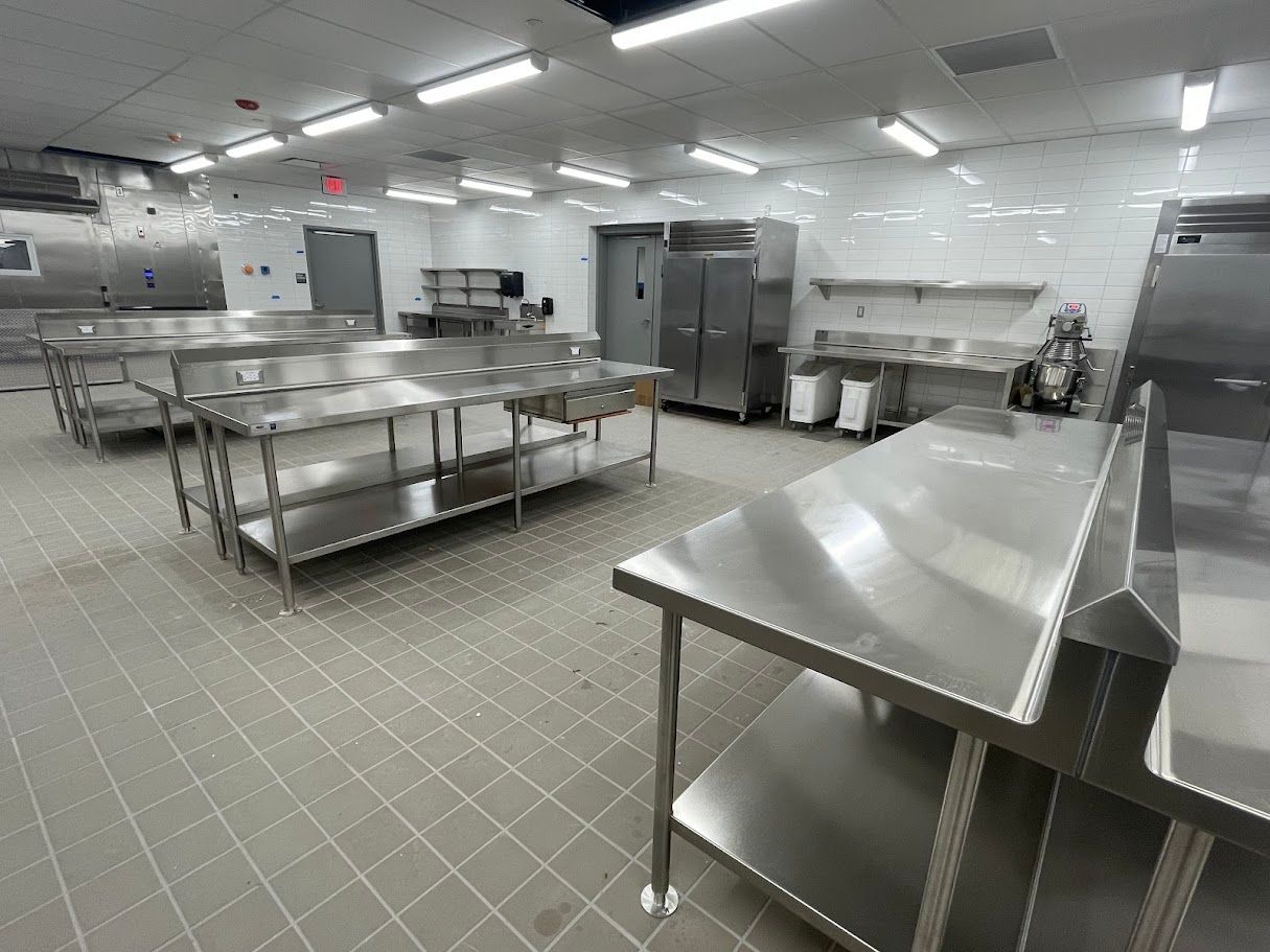 A clean, commercial kitchen with stainless steel prep tables, tile floors, and bright overhead lights.