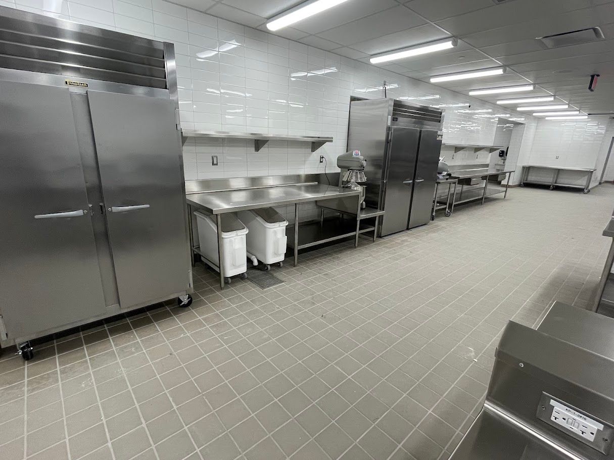 A wide, brightly lit industrial kitchen featuring stainless steel refrigerators, metal workstations, and tiled floors.