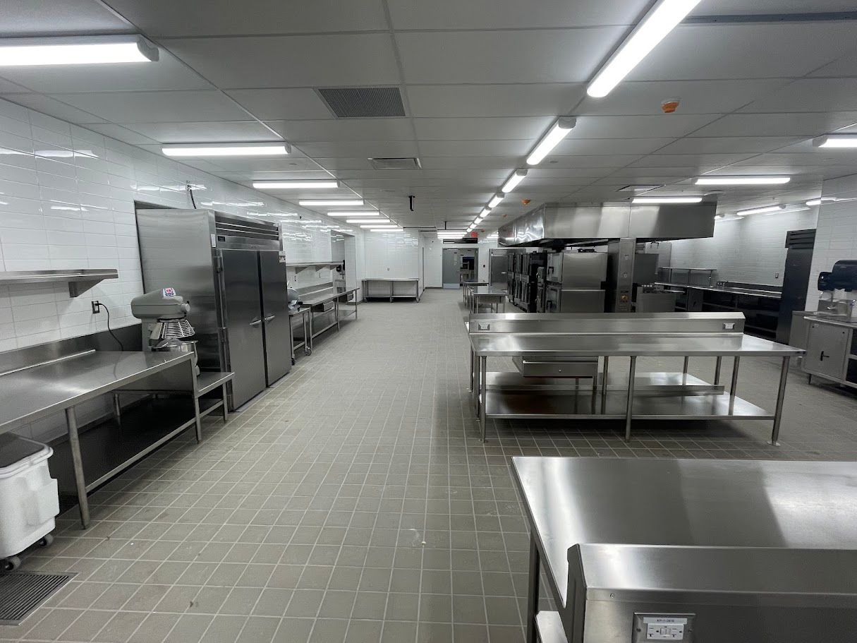 A wide-angle view of a commercial kitchen featuring stainless steel prep tables, industrial appliances, and tiled floors.