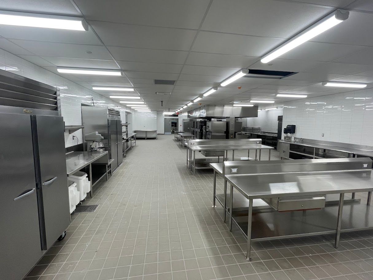 A commercial kitchen with stainless steel prep tables and refrigerators under bright overhead lights.