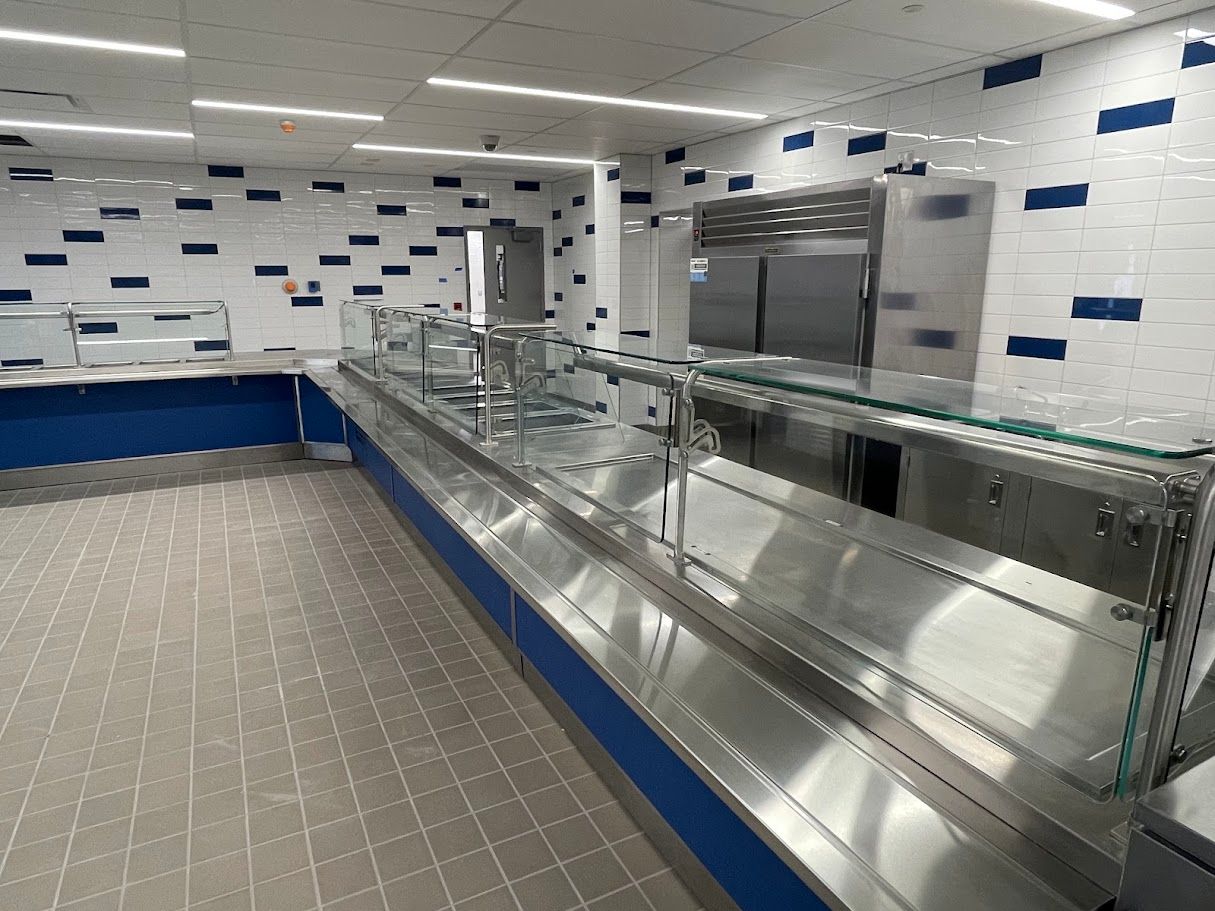 A stainless steel serving counter in an empty cafeteria with white tiled walls featuring blue decorative rectangles.