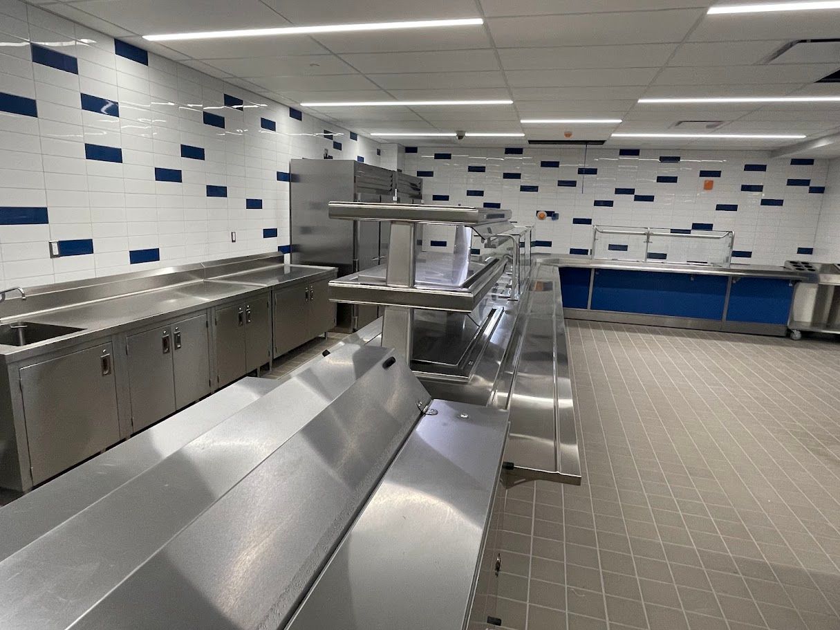 A stainless steel cafeteria serving line with tiled walls featuring blue accents in a brightly lit kitchen.
