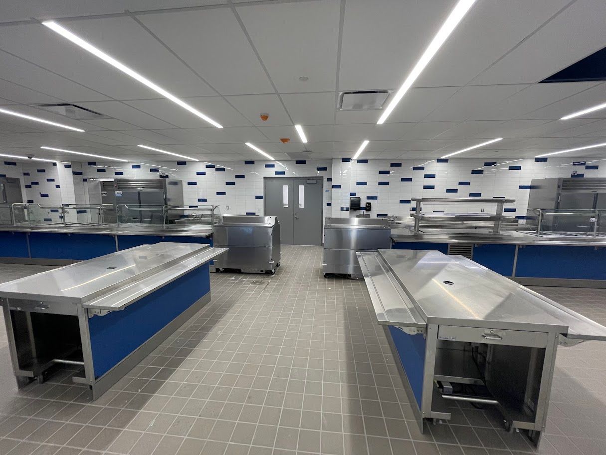 Empty industrial cafeteria with stainless steel serving counters, blue base panels, and a patterned tiled wall.