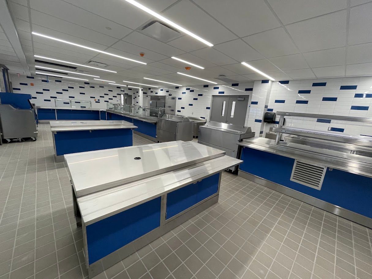 An empty school cafeteria with stainless steel food service counters, blue accents, white walls, and tiled floors.