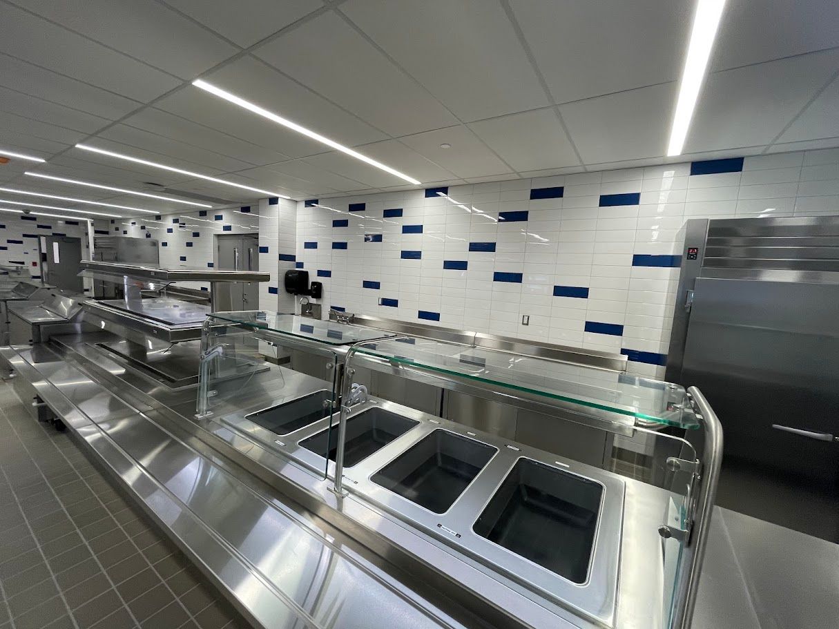 A modern, empty food serving station with stainless steel counters and a white wall with scattered blue rectangular tiles.