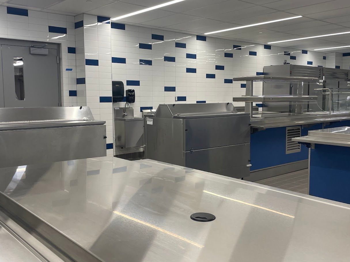 A commercial kitchen with stainless steel prep tables, metal equipment, and tiled walls with blue accent squares.