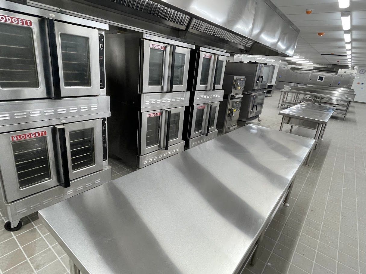 A commercial kitchen with rows of stainless steel ovens and prep tables on a tiled floor.