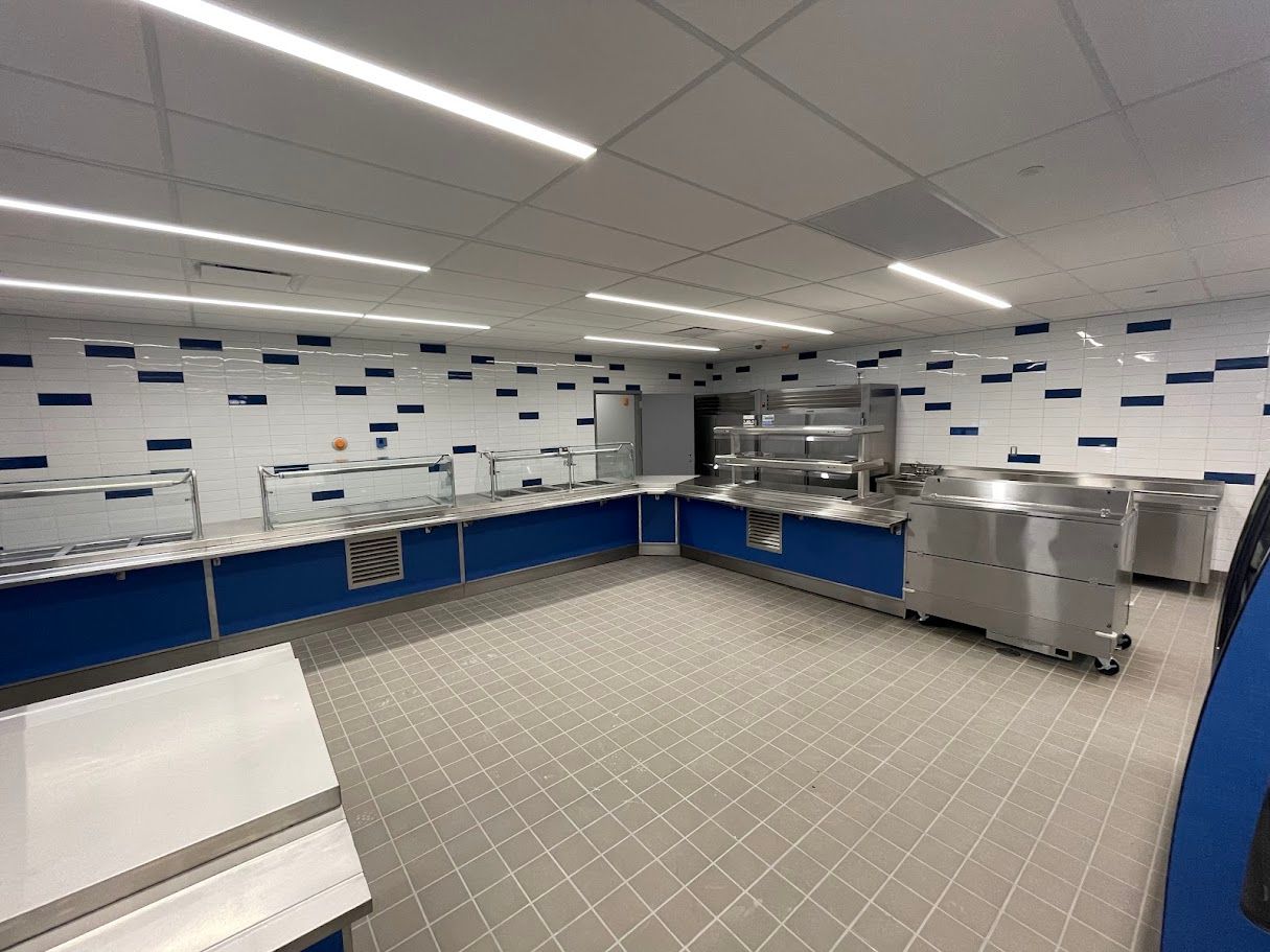 A bright, modern school cafeteria serving line with blue cabinets, white tiled walls, and stainless steel food stations.