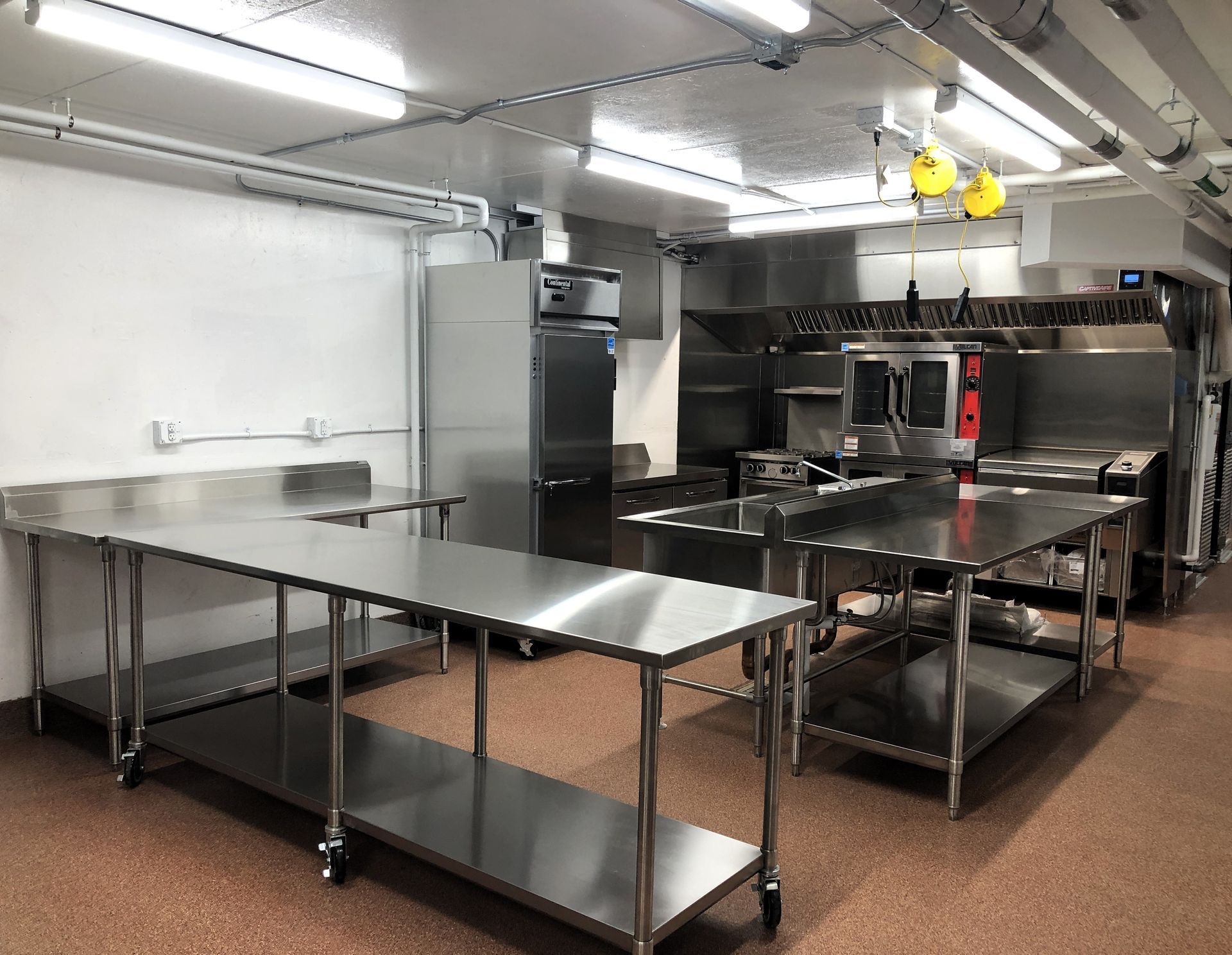 A bright commercial kitchen with stainless steel prep tables, a large refrigerator, and industrial appliances.