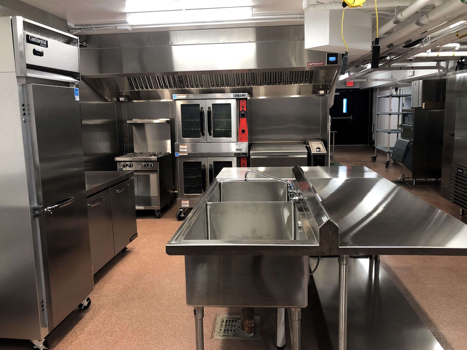 A modern, stainless steel commercial kitchen featuring a double-basin sink, oven units, and a refrigerator.