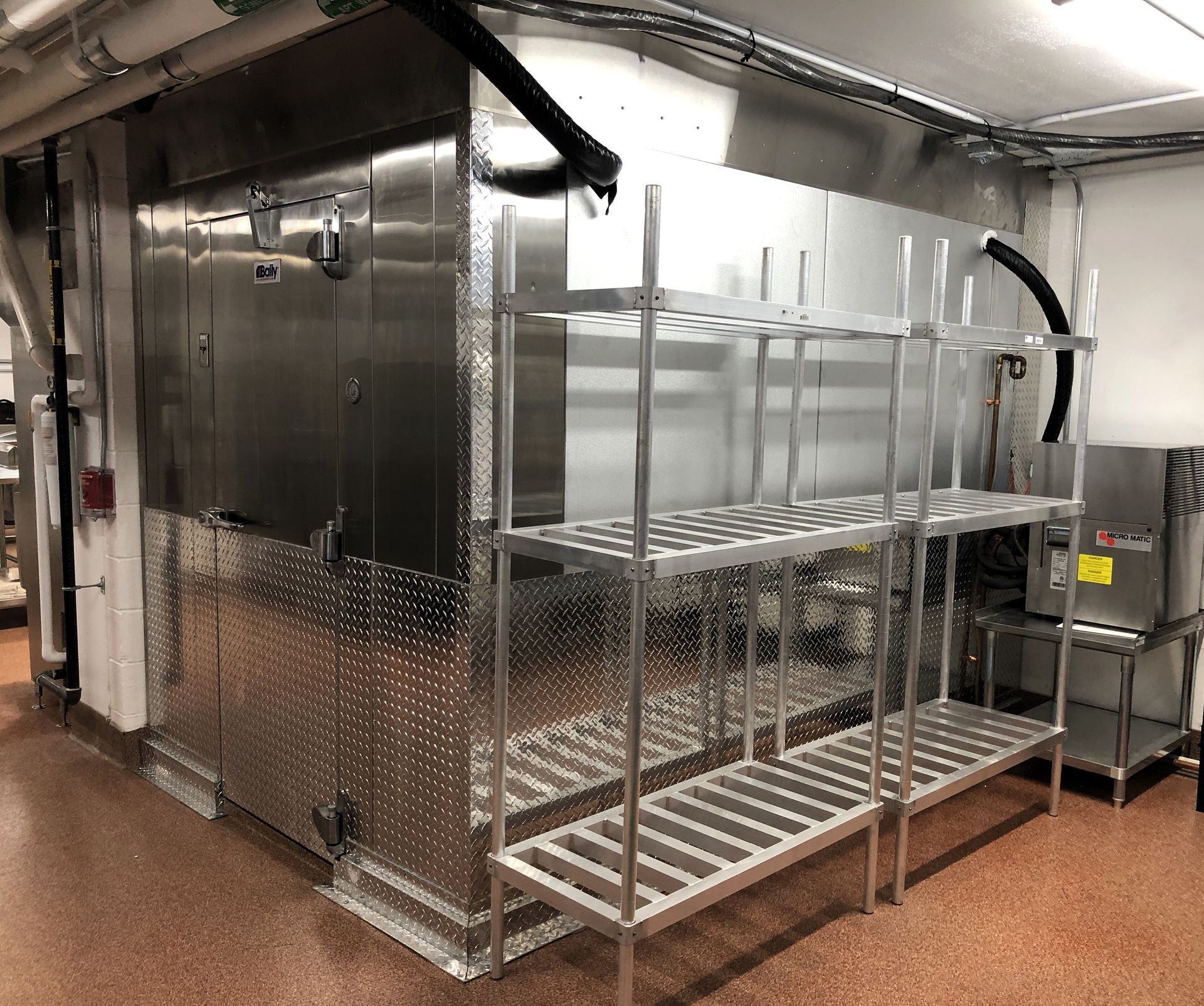 A metal walk-in cooler stands in a commercial room, with empty stainless steel shelving units in front of it.