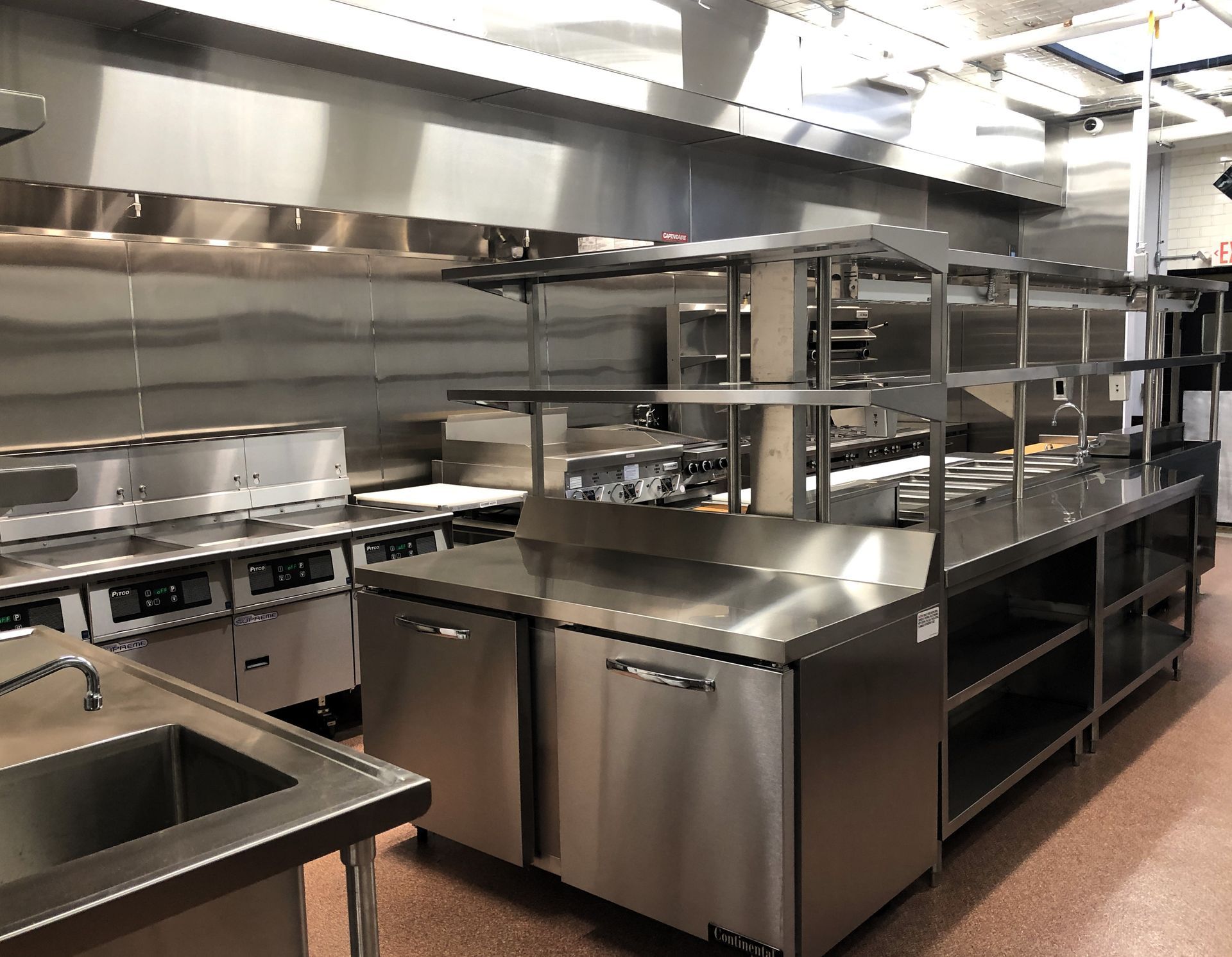 A modern commercial kitchen with stainless steel appliances, prep tables, and shelving under industrial ventilation hoods.