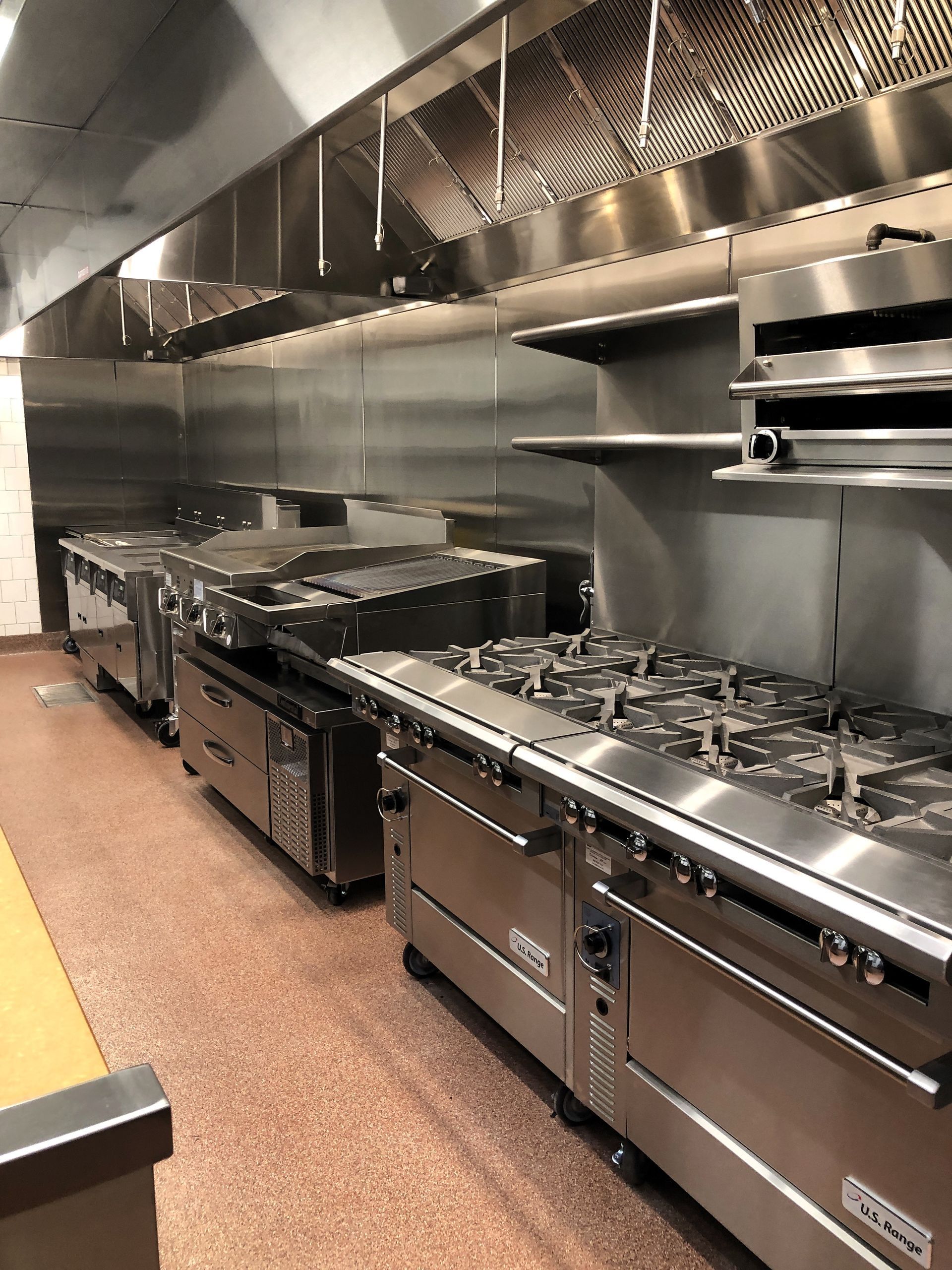 A commercial kitchen featuring a row of stainless steel stoves, ovens, and a metal exhaust hood against a tiled wall.