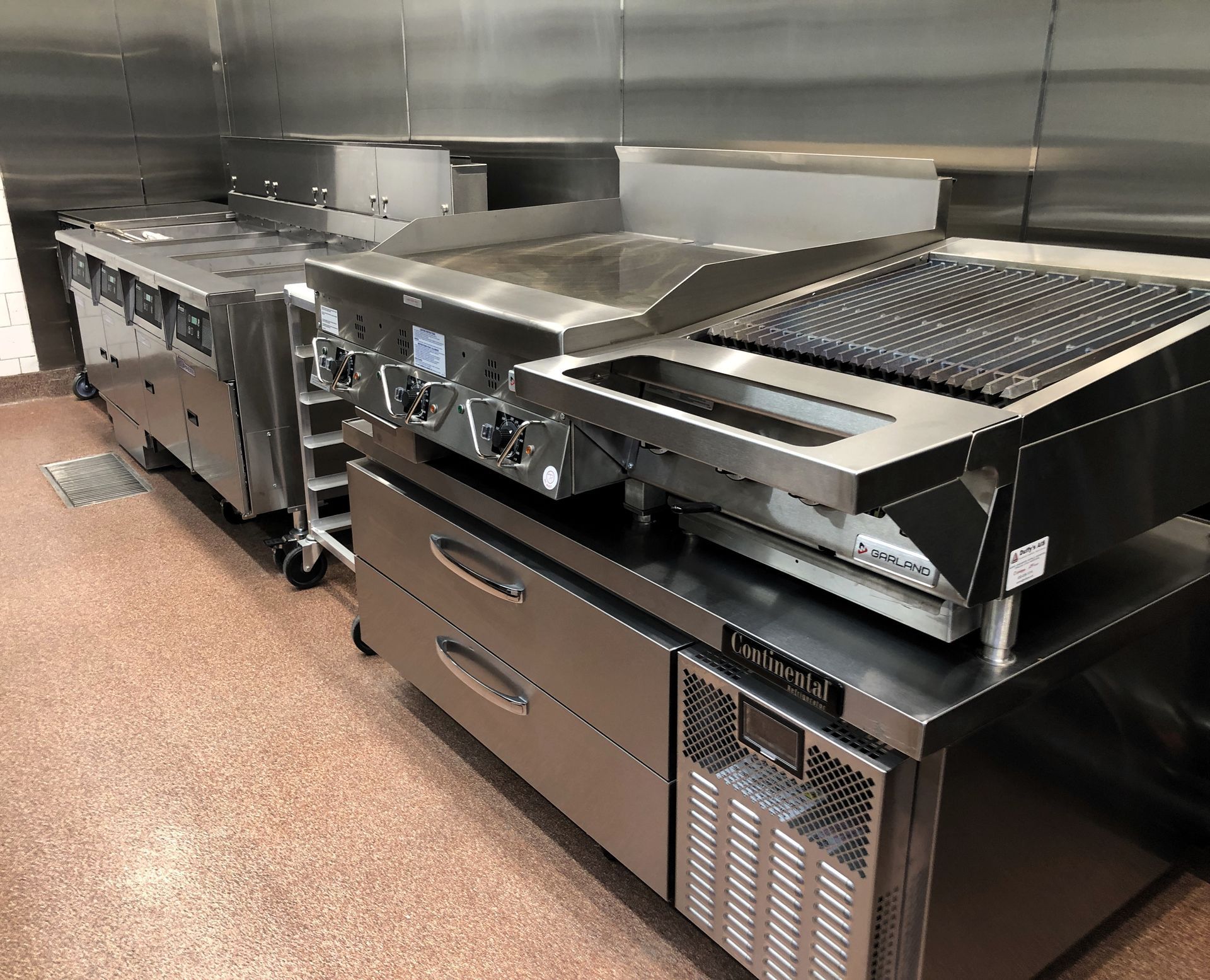 Stainless steel commercial kitchen cooking equipment, including a griddle and charbroiler, atop refrigerated drawers.