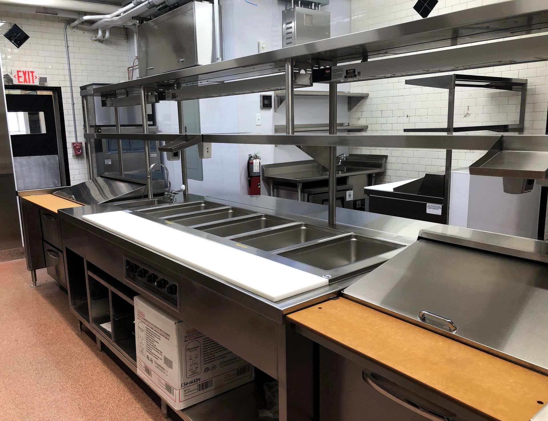 A commercial kitchen prep station with stainless steel counters, a food pan rail, a white cutting board, and metal shelving.