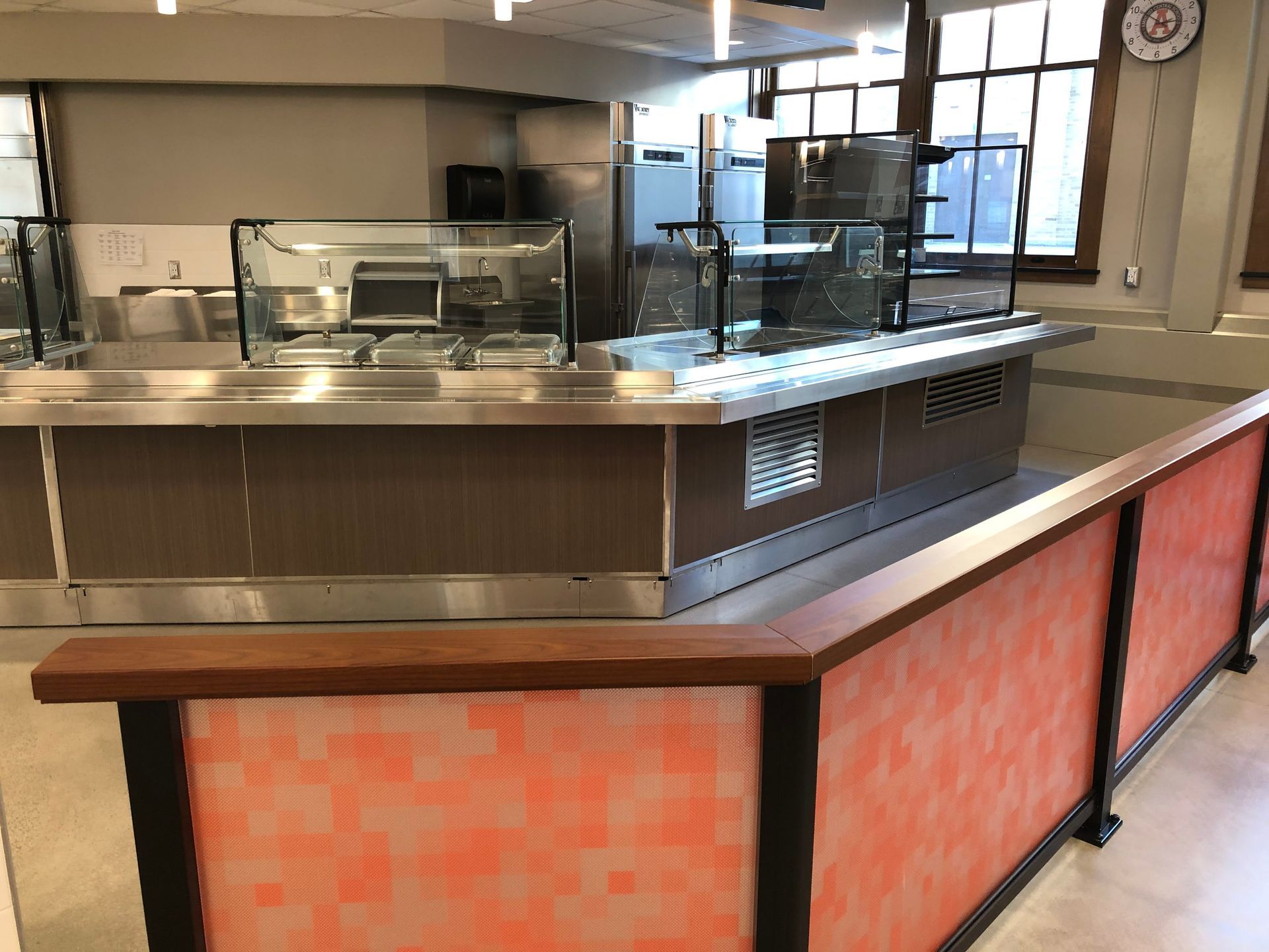 A modern cafeteria serving area with stainless steel counters, glass sneeze guards, and a decorative orange-paneled railing.