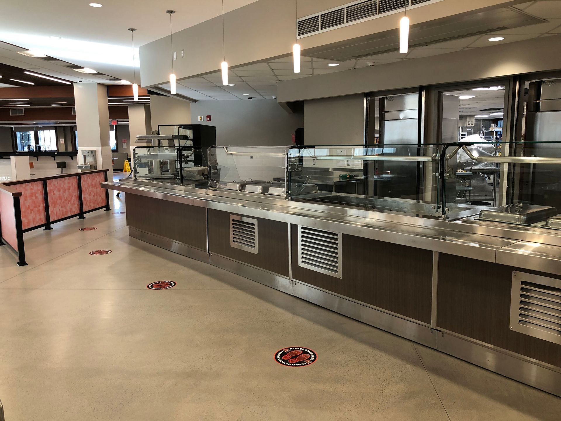 A long, empty cafeteria serving station with stainless steel counters, modern lighting, and social distancing floor markers.