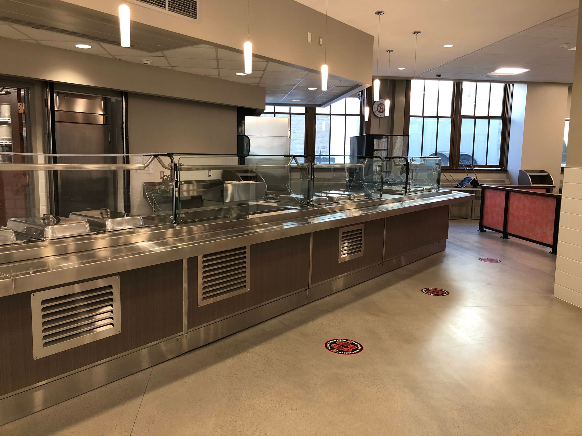 A modern cafeteria serving line with stainless steel counters, glass sneeze guards, and red floor decals.