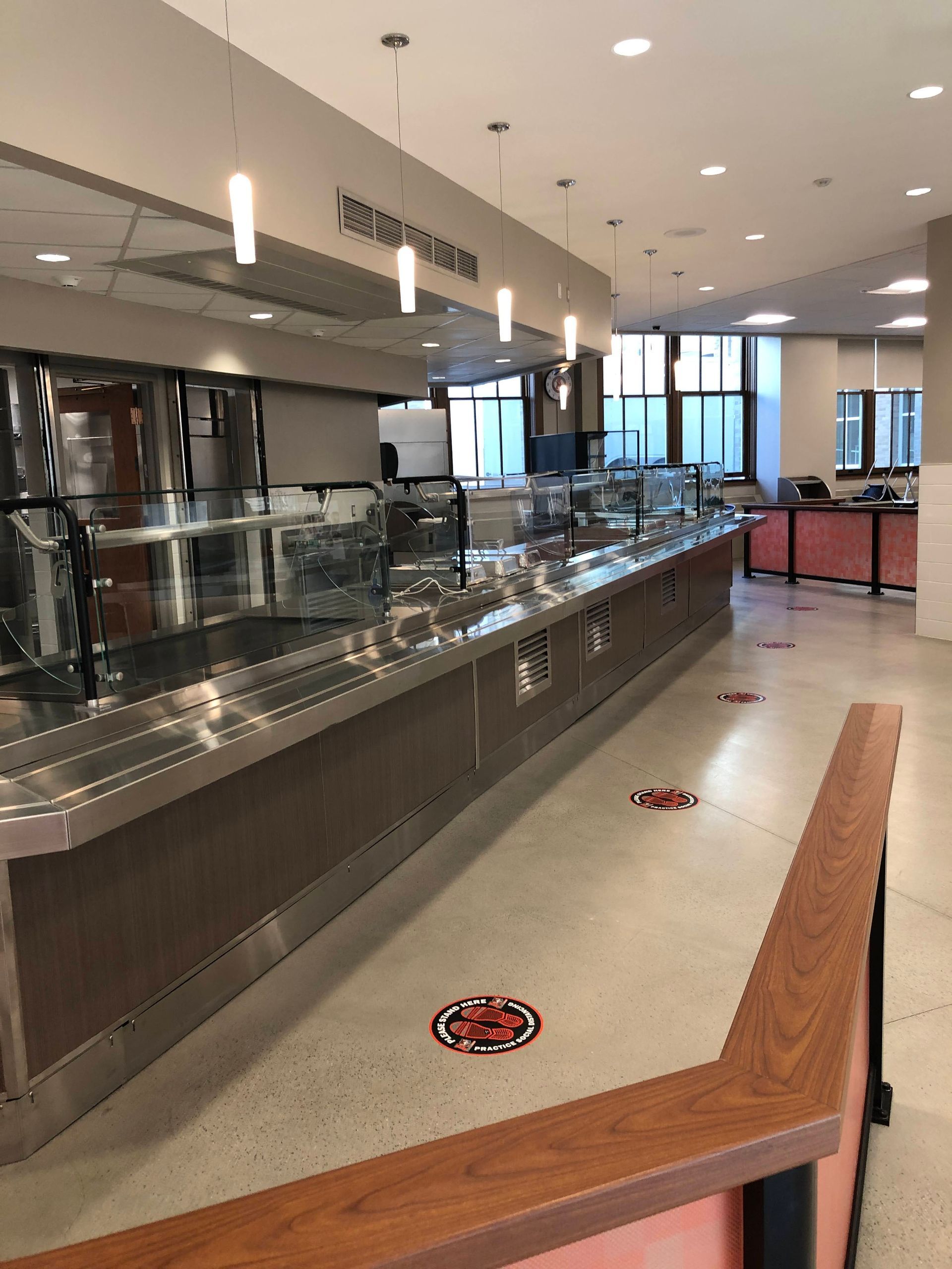 An empty cafeteria serving line with pendant lights and social distancing floor markers.