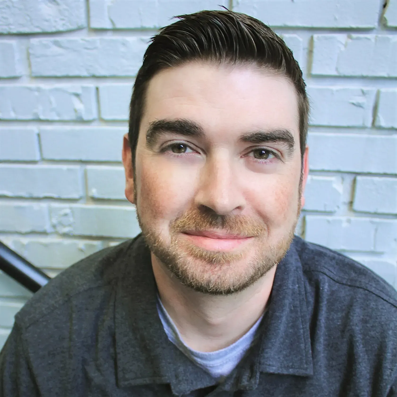 A person with short dark hair and facial hair wears a dark grey shirt in front of a white brick wall.