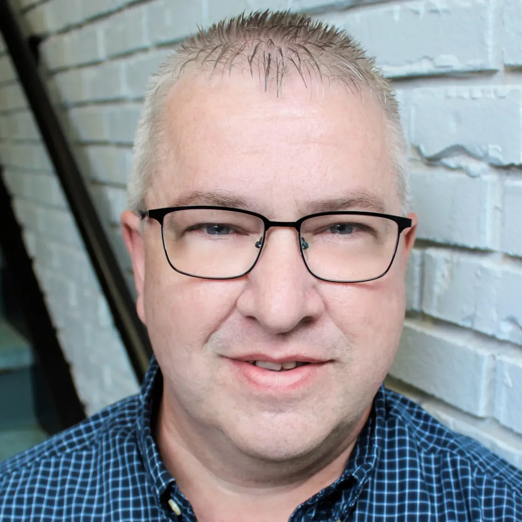 A person wearing glasses and a blue checkered shirt, smiling slightly while standing against a white brick wall.