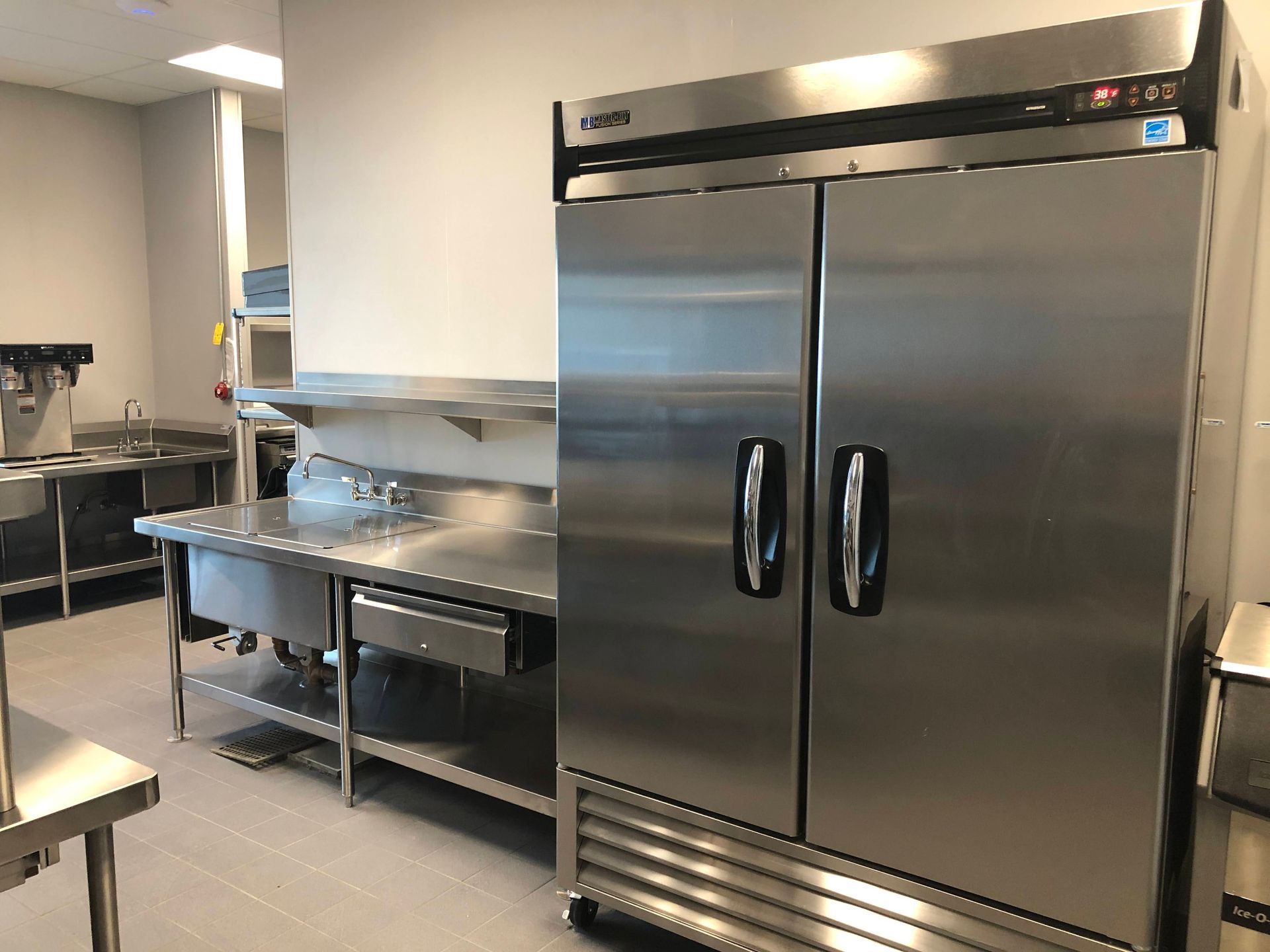 A commercial kitchen with a large stainless steel refrigerator next to a stainless steel prep table and sink.