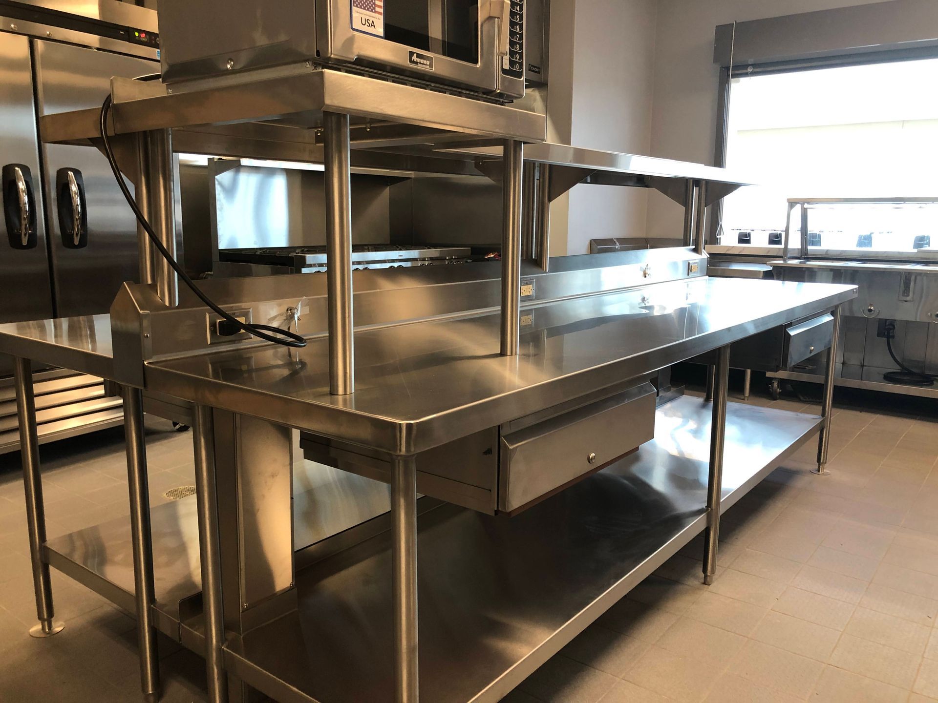 A stainless steel kitchen preparation table with an upper shelf and a microwave, positioned in a commercial kitchen.