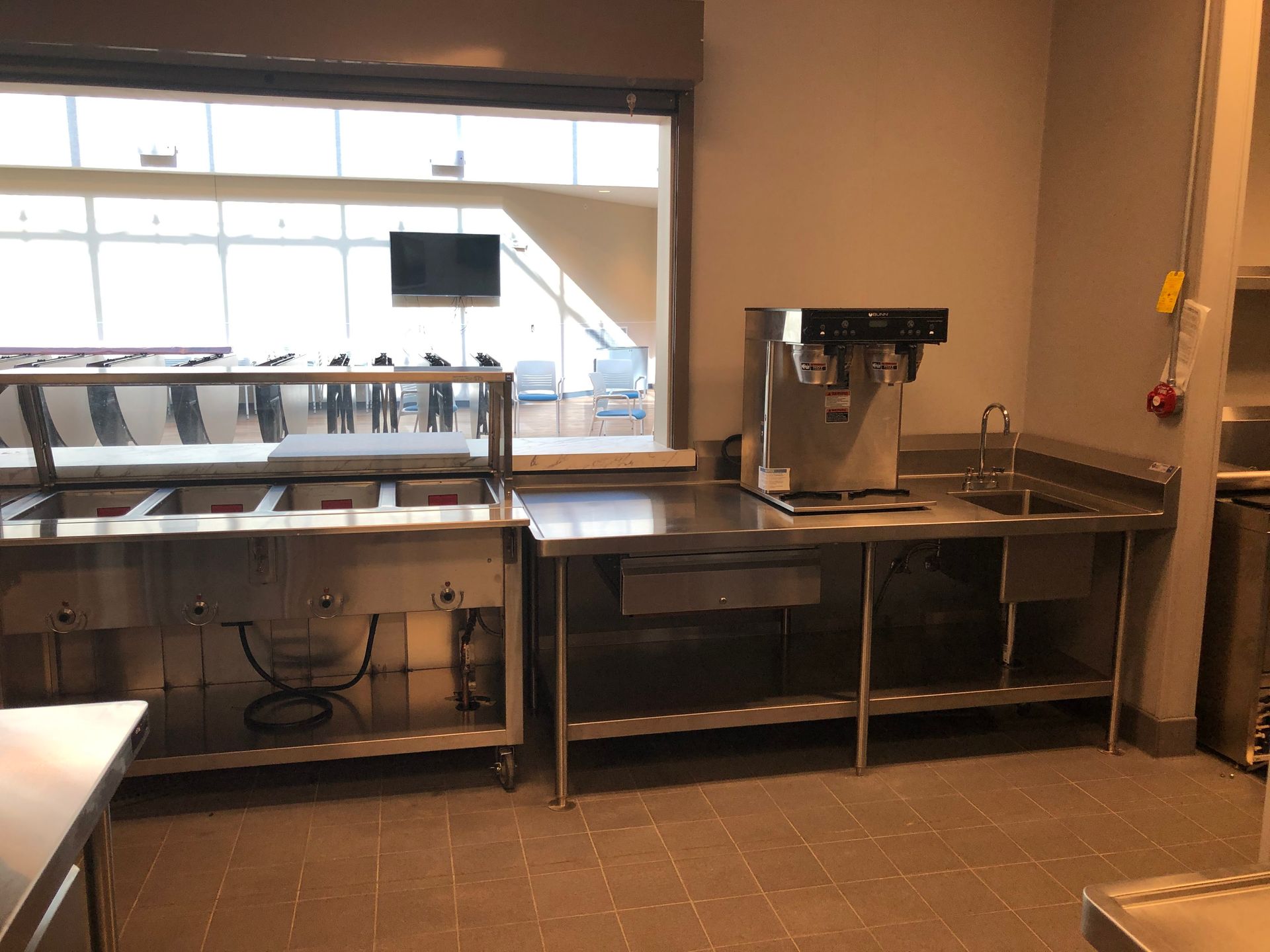 A commercial kitchen area featuring stainless steel counters, a food warmer, a coffee machine, and a sink by a window.