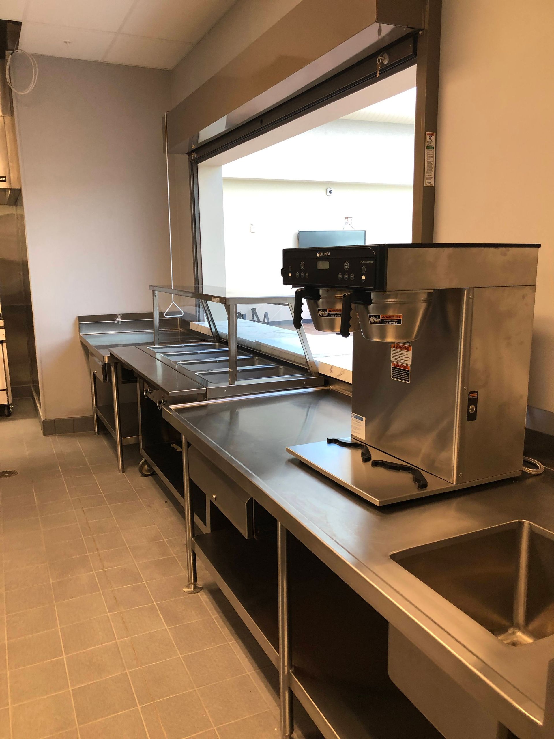 A stainless steel commercial coffee maker sits on a service counter in a kitchen next to a food prep station.