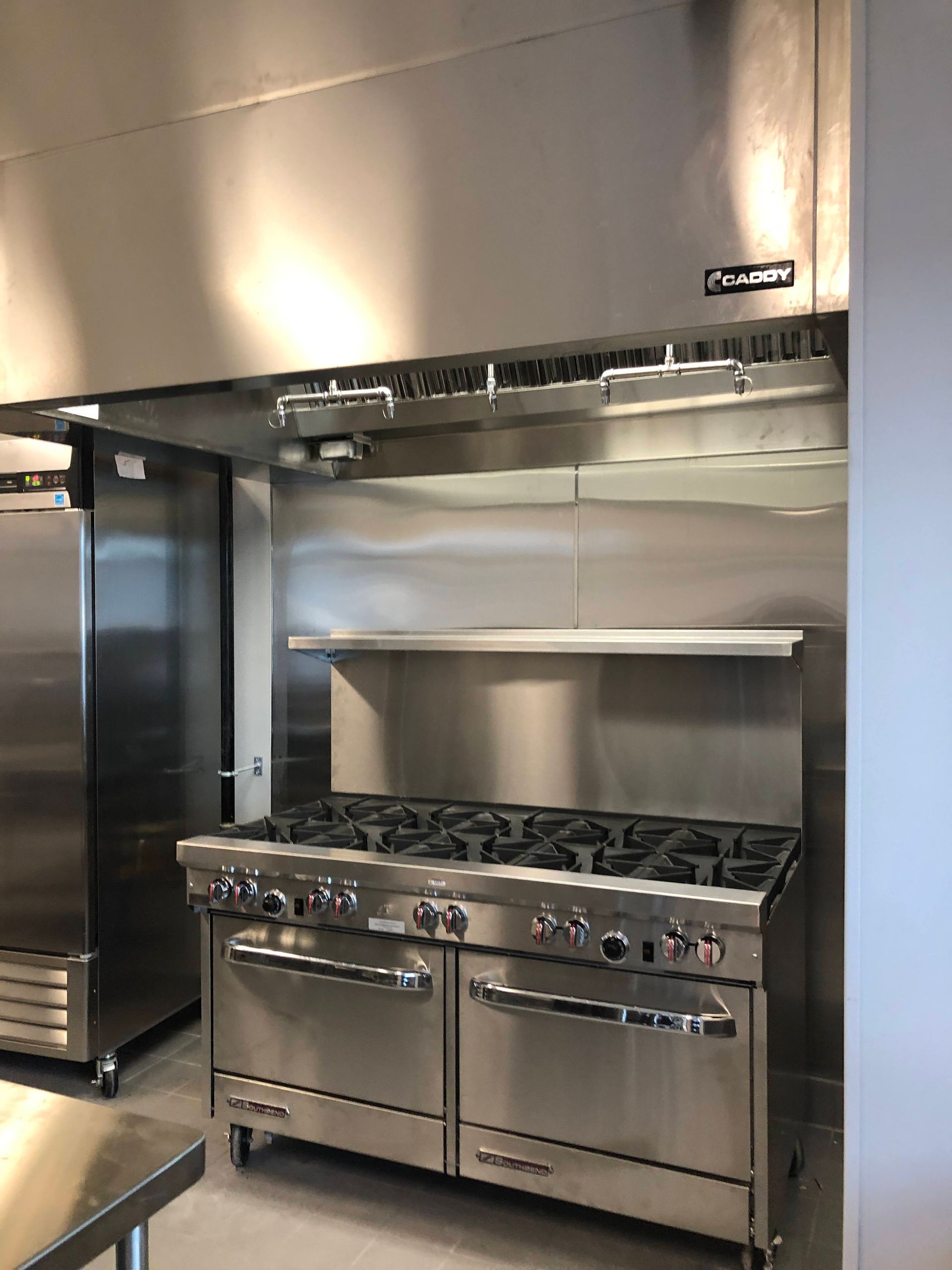 A stainless steel commercial range with six burners and two ovens sits beneath a large matching ventilation hood.
