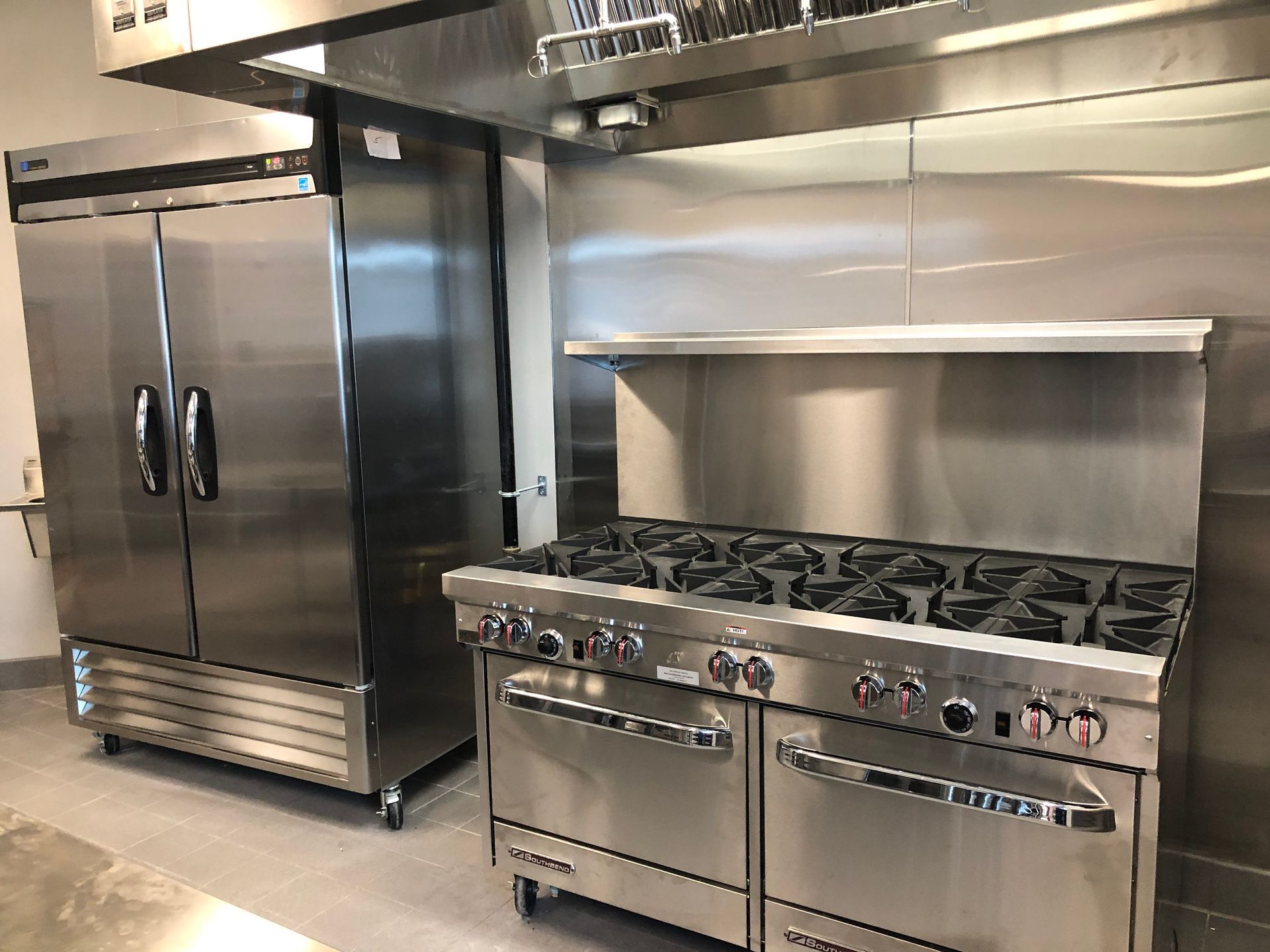 A commercial kitchen featuring a tall stainless steel refrigerator next to a large industrial gas stove under a hood.
